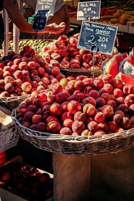 Baskets filled with ripe peaches are displayed at a market stall. Chalkboard signs indicate prices and origins of the fruits. A variety of produce can be seen in the background, with some partial view of people shopping.