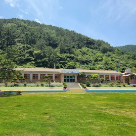A single-story building with a beige exterior and red-framed windows is situated against a backdrop of lush, green, forested hills. The building has a slightly elevated entrance with steps leading up to the main door. In front of the building, there is a large, well-manicured lawn with a few neatly trimmed bushes and a paved walkway lined with colorful stripes. The sky above is clear and blue, indicating a sunny day.