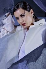 A person dressed in a white outfit is surrounded by sheer, flowing fabric. They have dark hair styled back and are wearing large, distinctive earrings. The lighting casts dramatic shadows on their face, highlighting their makeup and creating an artistic, fashion-forward atmosphere.