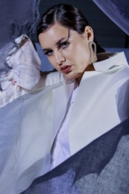 A person dressed in a white outfit is surrounded by sheer, flowing fabric. They have dark hair styled back and are wearing large, distinctive earrings. The lighting casts dramatic shadows on their face, highlighting their makeup and creating an artistic, fashion-forward atmosphere.