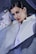 A person dressed in a white outfit is surrounded by sheer, flowing fabric. They have dark hair styled back and are wearing large, distinctive earrings. The lighting casts dramatic shadows on their face, highlighting their makeup and creating an artistic, fashion-forward atmosphere.