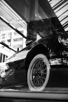 A black and white photograph of a classic vintage car with intricate wheel spokes and a shiny exterior. Reflections from windows and nearby architecture create an artsy, layered effect.