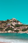 A hill overlooking the sea with multiple houses and villas nestled among lush greenery. The turquoise waters of the sea are in the foreground with small boats docked near the shoreline. The sky above is clear and bright, contributing to a serene and picturesque landscape.