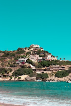 A hill overlooking the sea with multiple houses and villas nestled among lush greenery. The turquoise waters of the sea are in the foreground with small boats docked near the shoreline. The sky above is clear and bright, contributing to a serene and picturesque landscape.