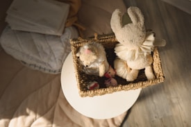 A woven basket on a white surface contains stuffed toys, including a plush rabbit with a ruffled collar and a bear. The background includes soft textiles like folded blankets or sheets placed on a surface.