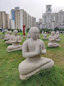 Multiple stone statues of meditating figures are arranged on a well-maintained green lawn, set against a backdrop of tall residential buildings. The statues are in a kneeling posture with hands clasped together, conveying a sense of tranquility and prayer.