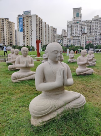 Multiple stone statues of meditating figures are arranged on a well-maintained green lawn, set against a backdrop of tall residential buildings. The statues are in a kneeling posture with hands clasped together, conveying a sense of tranquility and prayer.
