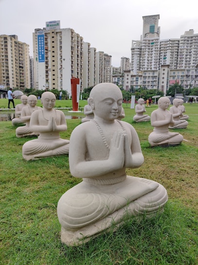 Multiple stone statues of meditating figures are arranged on a well-maintained green lawn, set against a backdrop of tall residential buildings. The statues are in a kneeling posture with hands clasped together, conveying a sense of tranquility and prayer.