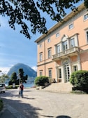 A grand villa with a peach-colored facade and large, elegant windows is situated near a serene lake, with lush green hills in the background. A path leads from the entrance, flanked by neatly trimmed hedges. A person is walking along the path, pushing a stroller. Overhanging branches partly frame the top of the scene.