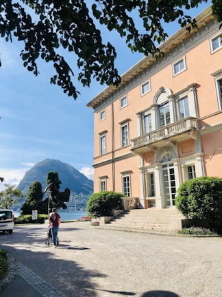 A grand villa with a peach-colored facade and large, elegant windows is situated near a serene lake, with lush green hills in the background. A path leads from the entrance, flanked by neatly trimmed hedges. A person is walking along the path, pushing a stroller. Overhanging branches partly frame the top of the scene.
