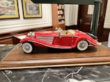 A detailed model of a vintage red convertible car with white accents is displayed on a wooden base. The car features intricate detailing, including chrome headlights, side mirrors, and wire-spoked wheels. The background includes decorative framed pictures and shelves, suggesting an indoor setting.