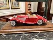 A detailed model of a vintage red convertible car with white accents is displayed on a wooden base. The car features intricate detailing, including chrome headlights, side mirrors, and wire-spoked wheels. The background includes decorative framed pictures and shelves, suggesting an indoor setting.