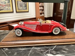 A detailed model of a vintage red convertible car with white accents is displayed on a wooden base. The car features intricate detailing, including chrome headlights, side mirrors, and wire-spoked wheels. The background includes decorative framed pictures and shelves, suggesting an indoor setting.