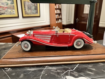 A detailed model of a vintage red convertible car with white accents is displayed on a wooden base. The car features intricate detailing, including chrome headlights, side mirrors, and wire-spoked wheels. The background includes decorative framed pictures and shelves, suggesting an indoor setting.