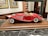 A detailed model of a vintage red convertible car with white accents is displayed on a wooden base. The car features intricate detailing, including chrome headlights, side mirrors, and wire-spoked wheels. The background includes decorative framed pictures and shelves, suggesting an indoor setting.
