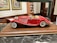 A detailed model of a vintage red convertible car with white accents is displayed on a wooden base. The car features intricate detailing, including chrome headlights, side mirrors, and wire-spoked wheels. The background includes decorative framed pictures and shelves, suggesting an indoor setting.