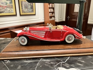 A detailed model of a vintage red convertible car with white accents is displayed on a wooden base. The car features intricate detailing, including chrome headlights, side mirrors, and wire-spoked wheels. The background includes decorative framed pictures and shelves, suggesting an indoor setting.