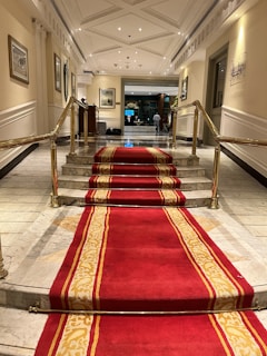 A luxurious indoor setting featuring a marble staircase with a red and gold carpet runner. The staircase is flanked by polished brass handrails and leads to a well-lit hallway. The hallway has framed pictures hanging on the walls and a person is visible at a distance.. The ceiling is intricately designed, and there are bright lights illuminating the area.