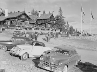 A black and white photograph captures a scene outside a large, rustic wooden lodge surrounded by pine trees, with multiple vintage cars parked in the foreground. Several people are gathered near the lodge, some standing and others walking, suggesting a social event or gathering. Flags are flying on tall poles on the right side of the image.