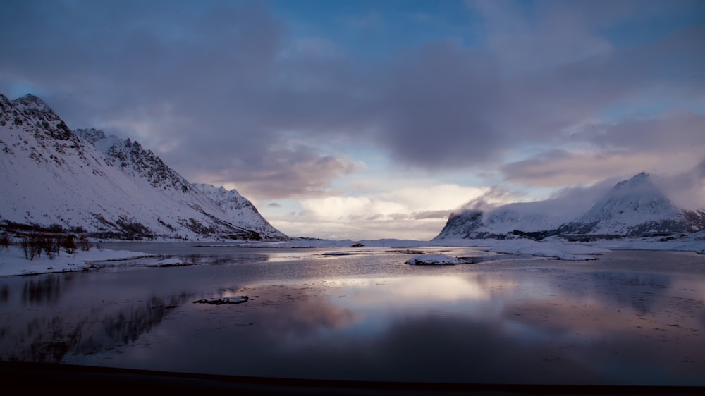 a lake surrounded by snow covered mountains under a cloudy sky