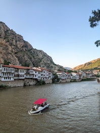 A scenic riverside view features traditional Ottoman-style houses with red-tiled roofs, nestled against a backdrop of rocky hills. A small motorboat with a red canopy glides along the wide river, carrying a few passengers. The sky is clear, and a cluster of trees is partially visible on the right.