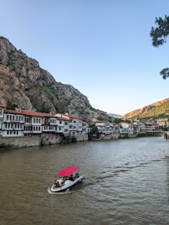 A scenic riverside view features traditional Ottoman-style houses with red-tiled roofs, nestled against a backdrop of rocky hills. A small motorboat with a red canopy glides along the wide river, carrying a few passengers. The sky is clear, and a cluster of trees is partially visible on the right.