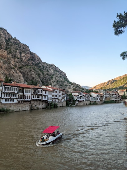 A scenic riverside view features traditional Ottoman-style houses with red-tiled roofs, nestled against a backdrop of rocky hills. A small motorboat with a red canopy glides along the wide river, carrying a few passengers. The sky is clear, and a cluster of trees is partially visible on the right.