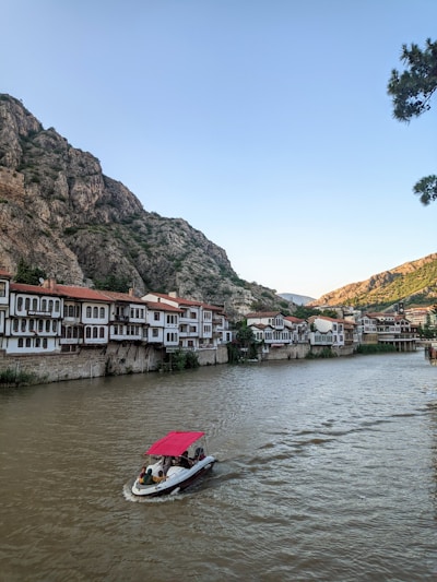 A scenic riverside view features traditional Ottoman-style houses with red-tiled roofs, nestled against a backdrop of rocky hills. A small motorboat with a red canopy glides along the wide river, carrying a few passengers. The sky is clear, and a cluster of trees is partially visible on the right.