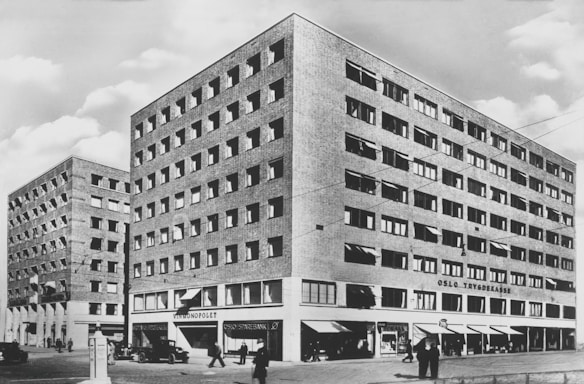 A black and white photograph of a mid-20th century cityscape featuring two large brick buildings with multiple rows of windows. The building on the right has 'Oslo Trygdekasse' written on it. Below, there are shopfronts with signage including 'Vinmonopolet' and 'Oslo Sparebank'. Several pedestrians are walking on the sidewalk, and vintage cars are parked along the street.