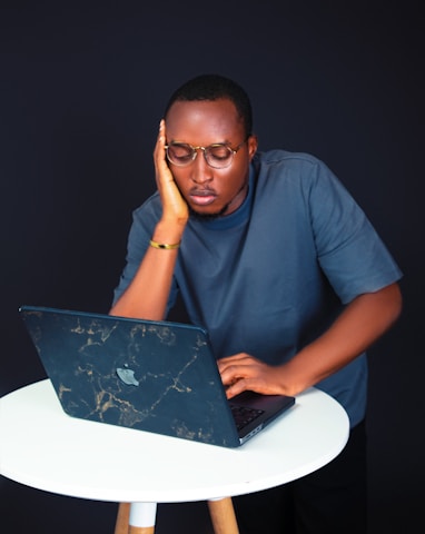 A man with glasses is leaning on a round white table with one hand resting on his cheek and the other typing on a laptop. He appears focused and deep in thought, dressed in a casual dark t-shirt. The background is dark, creating a contrast with the white table and his attire.