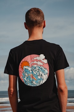 A person with short hair is standing with their back to the camera, wearing a black t-shirt featuring a vibrant graphic design. The design on the shirt is of a stylized wave, a sun, and clouds, rendered in a cartoon-like style with a mix of pink, blue, and orange colors. The scene is set against an outdoor backdrop with a view of the ocean and a clear sky, creating a calm and relaxed atmosphere.