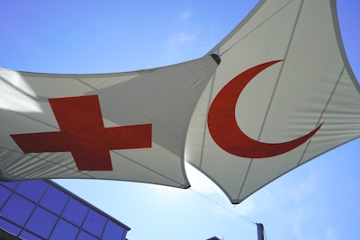Two triangular white canopies are displayed, each bearing prominent red symbols: a cross and a crescent. The canopies are suspended against a clear blue sky, with a corner of a modern glass building visible below.