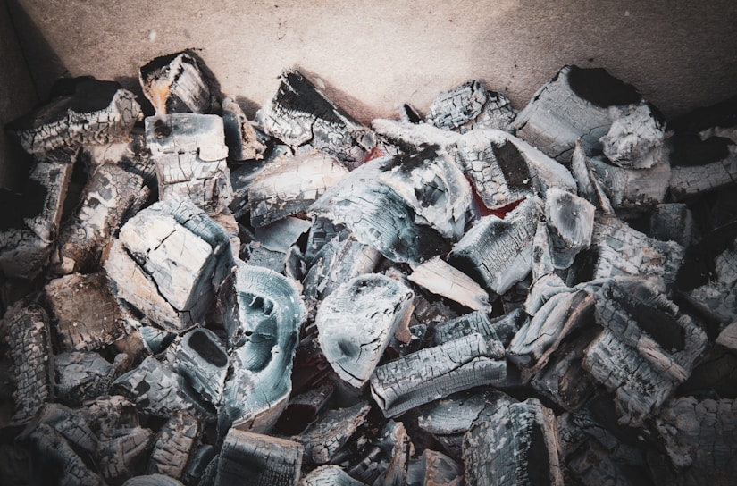 A collection of gray charcoal fragments and ashes, possibly remnants of a fire. The chunks appear irregular and layered, with sharp edges and varying shades of gray and black. The texture is rough, and some pieces have a slightly white ash covering.