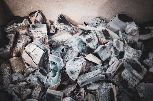 A collection of gray charcoal fragments and ashes, possibly remnants of a fire. The chunks appear irregular and layered, with sharp edges and varying shades of gray and black. The texture is rough, and some pieces have a slightly white ash covering.
