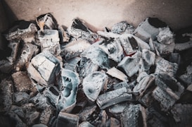 A collection of gray charcoal fragments and ashes, possibly remnants of a fire. The chunks appear irregular and layered, with sharp edges and varying shades of gray and black. The texture is rough, and some pieces have a slightly white ash covering.