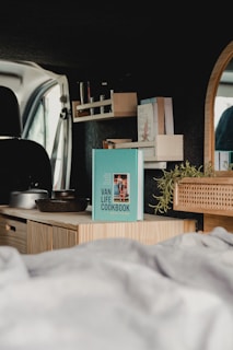 A cozy van interior features a bookshelf with a small collection of books, including a prominent blue cookbook titled 'Van Life Cookbook.' A wooden countertop displays kitchenware and a potted plant, adding a touch of greenery. A round mirror is partially visible, reflecting the van's window.