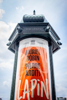 A large cylindrical advertising column with the word 'Theatres' embossed at the top. Below, a poster features the names 'Muriel Robin' and 'Pierre Arditi' along with the title 'Lapin' in bold, bright orange lettering. The design has a vintage look with detailed ornate decorations at the top.