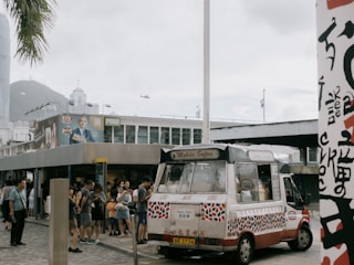 An ice cream van is parked on a city street, attracting a line of people waiting to be served. The van is decorated with colorful patterns and has signage indicating 'Mobile Softee.' Nearby is a billboard featuring a man, with a helicopter flying in the background. The setting appears urban with modern buildings and a small crowd gathered.
