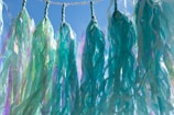 Multiple strands of translucent, colorful streamers are loosely tied to a rope, hanging against a clear blue sky. The streamers display a mix of iridescent green and blue hues, creating a whimsical and vibrant effect as they sway.