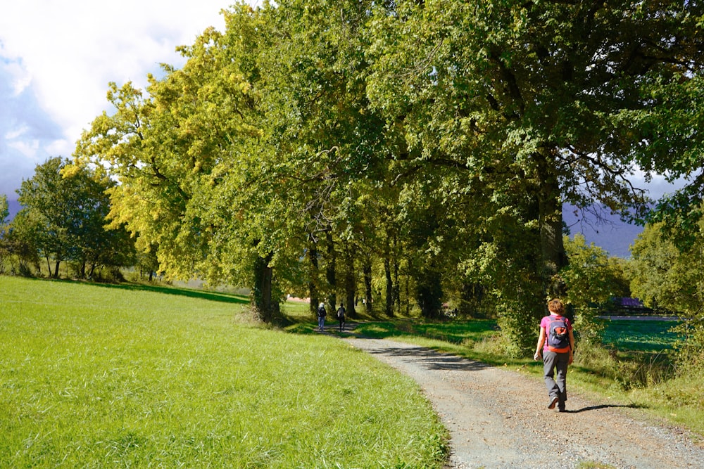 a woman walking down a dirt road next to a lush green field