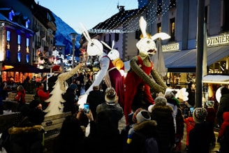 A festive street scene with giant animal puppets dressed in colorful costumes parading through a crowd of people. The setting is a charming town with snow-covered rooftops and mountains in the background. Strings of decorative lights illuminate the street, enhancing the vibrant atmosphere. Onlookers, some with children, are captured taking photographs and enjoying the spectacle.