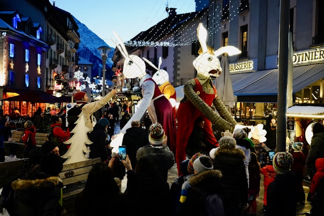 A festive street scene with giant animal puppets dressed in colorful costumes parading through a crowd of people. The setting is a charming town with snow-covered rooftops and mountains in the background. Strings of decorative lights illuminate the street, enhancing the vibrant atmosphere. Onlookers, some with children, are captured taking photographs and enjoying the spectacle.