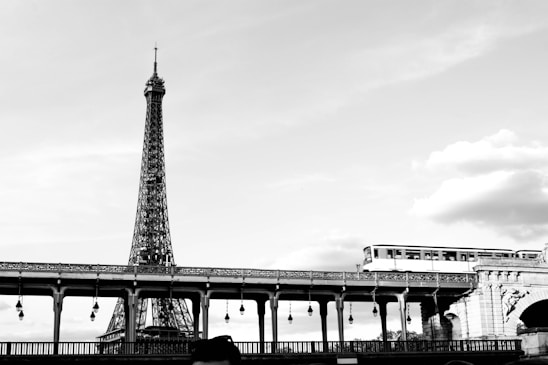 A black and white photograph featuring the iconic Eiffel Tower in the background and a bridge in the foreground with a metro train crossing over it. The sky is partially cloudy, adding some texture to the otherwise clean and minimalist scene.