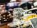 A detailed view of an intricate car engine model displayed on a transparent stand, with a blurred race car in the background. The scene suggests a motorsport or automotive theme.