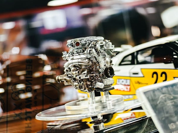 A detailed view of an intricate car engine model displayed on a transparent stand, with a blurred race car in the background. The scene suggests a motorsport or automotive theme.
