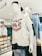 A mannequin wearing a white T-shirt with 'Brillo' written in red and blue text, combined with a pair of blue jeans. The mannequin is also wearing a black baseball cap backwards and white over-ear headphones. Behind it, there's another mannequin with glasses and a shirt, surrounded by neatly folded clothes on shelves.