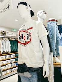 A mannequin wearing a white T-shirt with 'Brillo' written in red and blue text, combined with a pair of blue jeans. The mannequin is also wearing a black baseball cap backwards and white over-ear headphones. Behind it, there's another mannequin with glasses and a shirt, surrounded by neatly folded clothes on shelves.