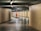 A person in bright orange work attire is sweeping the floor in a tiled underground passageway. The area is dimly lit with rows of overhead lights, creating a serene and focused atmosphere. Another person is walking away in the background, adding depth to the scene.