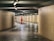 A person in bright orange work attire is sweeping the floor in a tiled underground passageway. The area is dimly lit with rows of overhead lights, creating a serene and focused atmosphere. Another person is walking away in the background, adding depth to the scene.