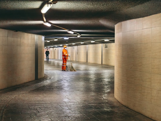 A person in bright orange work attire is sweeping the floor in a tiled underground passageway. The area is dimly lit with rows of overhead lights, creating a serene and focused atmosphere. Another person is walking away in the background, adding depth to the scene.